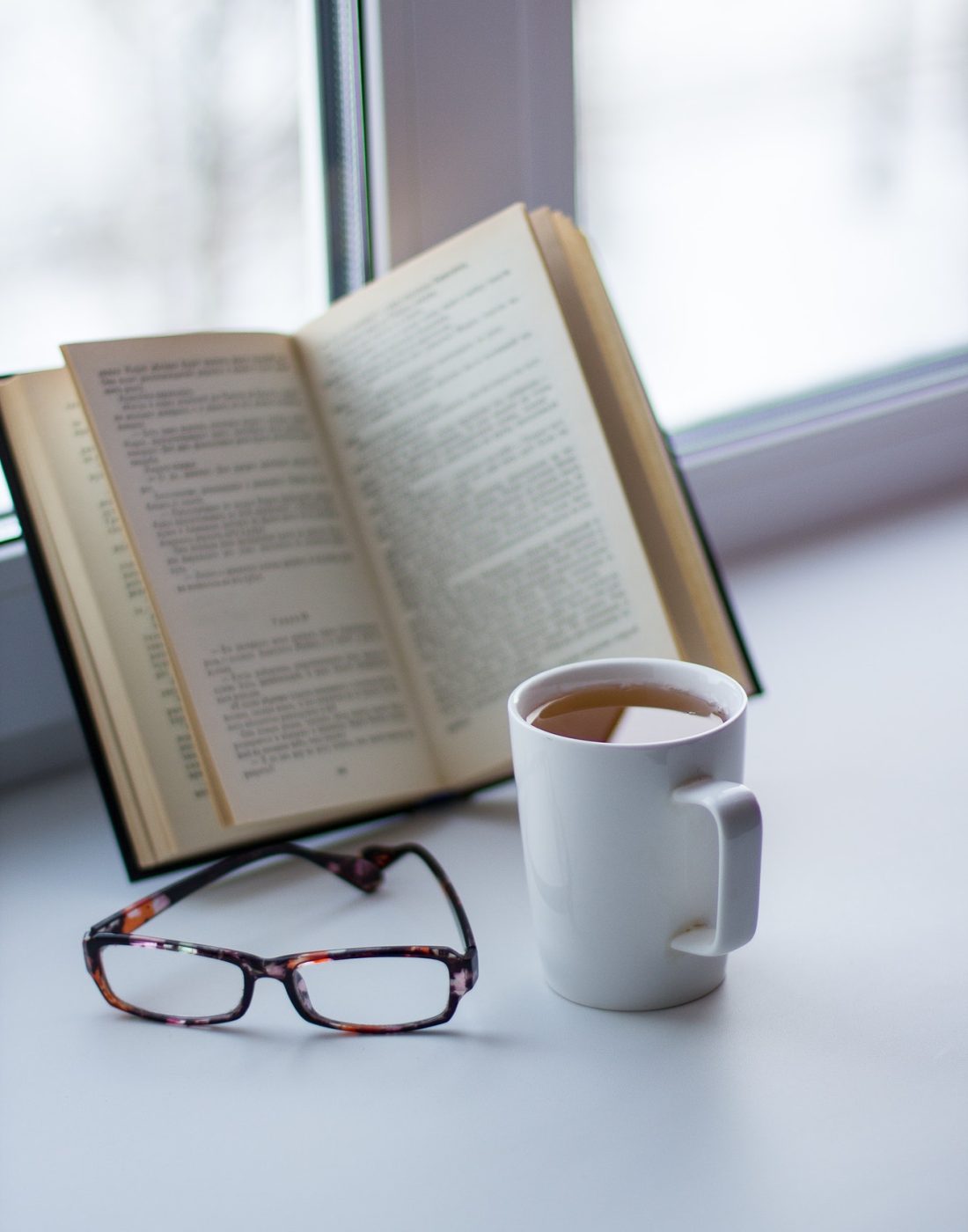 Open book, glasses, and coffee cup on a white table by the window.